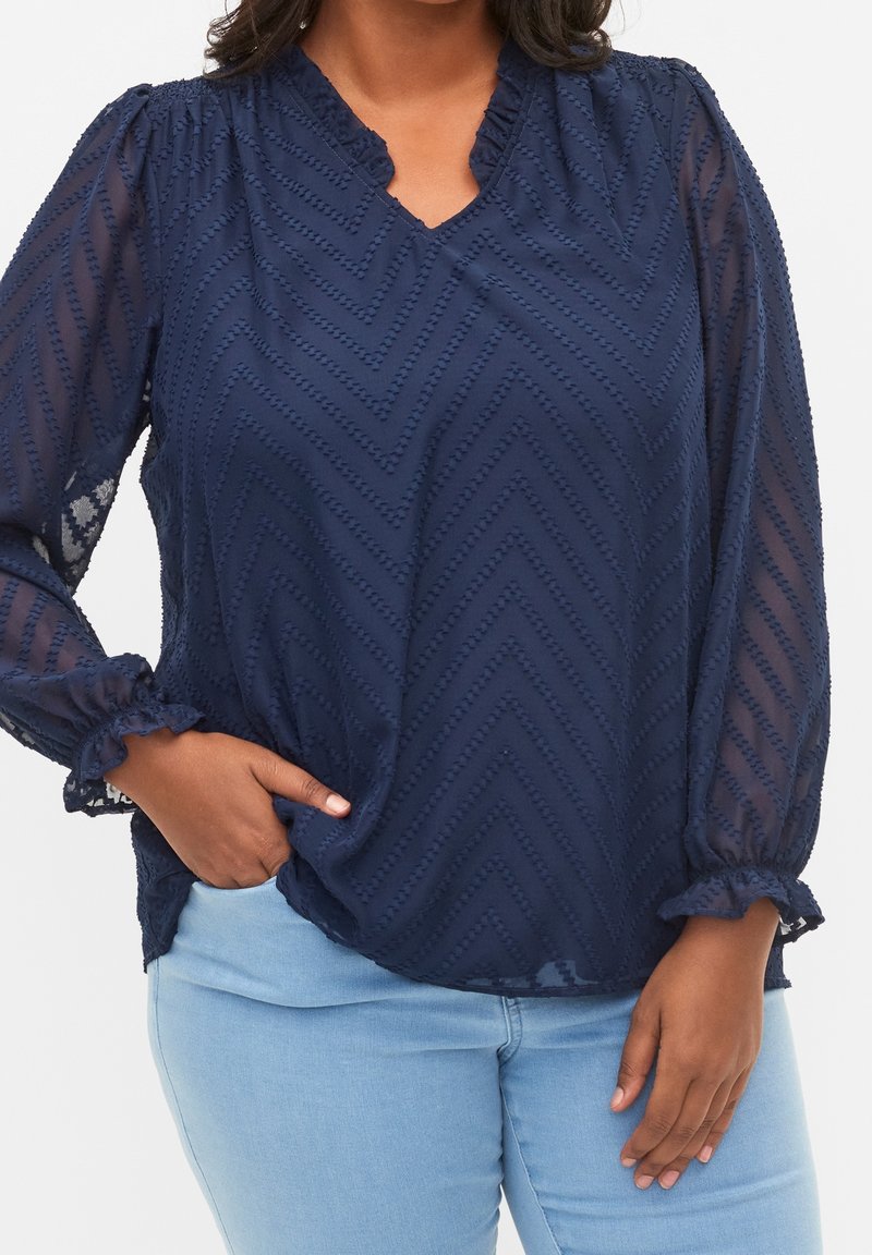 Femme portant un chemisier texturé bleu marine avec des manches longues transparentes et un jean bleu clair, une main dans la poche et l'autre reposant sur la cuisse.