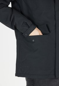 Sort vandtæt jakke med en struktureret finish, der har en synlig knaplomme på brystet og brunet ærmedetalje.