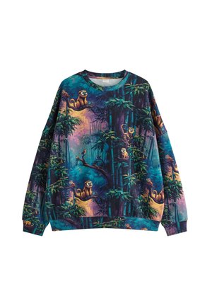 Ung mand med kort blondt hår iført farverig jungle-tema sweatshirt, lyseblå jeans og beige sneakers, står på ensfarvet baggrund.