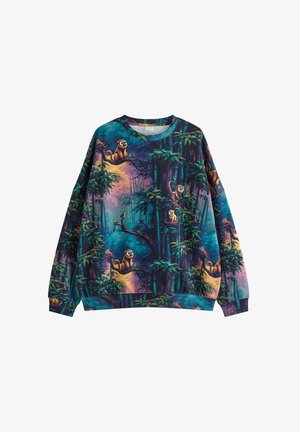 Jonge man met kort blond haar draagt kleurrijke sweatshirt met junglethema, lichtblauwe jeans en beige sneakers, staand voor een effen achtergrond.