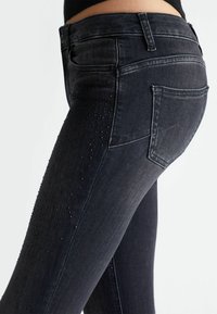 Jeans de denim oscuro con un ajuste slim, con un ligero brillo y acentos decorativos de tachuelas a lo largo de los lados. Bolsillos traseros clásicos, cierre en la cintura visible.