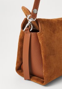 Brun ruskind og læder håndtaske med sølvfarvet hardware, afrundede kanter og en kontrastfuld tekstureret finish. Justerbar strop øverst.