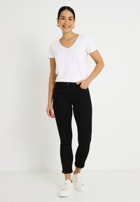 Jeans ajustados negros con bolsillos delanteros, combinados con una camiseta blanca de cuello en V y zapatillas blancas. El conjunto presenta un diseño casual y detalles sutiles.