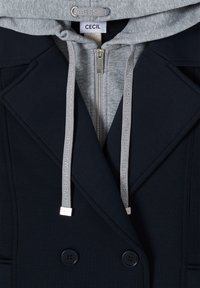 Manteau bleu marine à double boutonnage porté sur un sweat à capuche gris clair avec cordons arborant l'inscription « Découvrez de nouveaux endroits » et une fermeture éclair centrale.