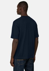 Homme portant un t-shirt bleu marine uni à manches courtes et un jean en denim foncé, vu de dos sur un fond neutre.