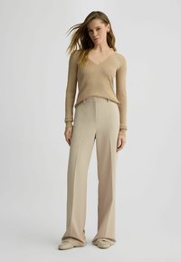 Top lungo a maniche lunghe beige a costine con scollatura a V, abbinato a pantaloni beige a vita alta e gamba larga. Il modello indossa scarpe slip-on beige morbide.