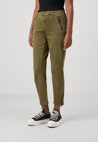 Olive green cargo pants with zippered front pockets, tapered leg, and ankle zippers. Worn with black sneakers featuring white laces.