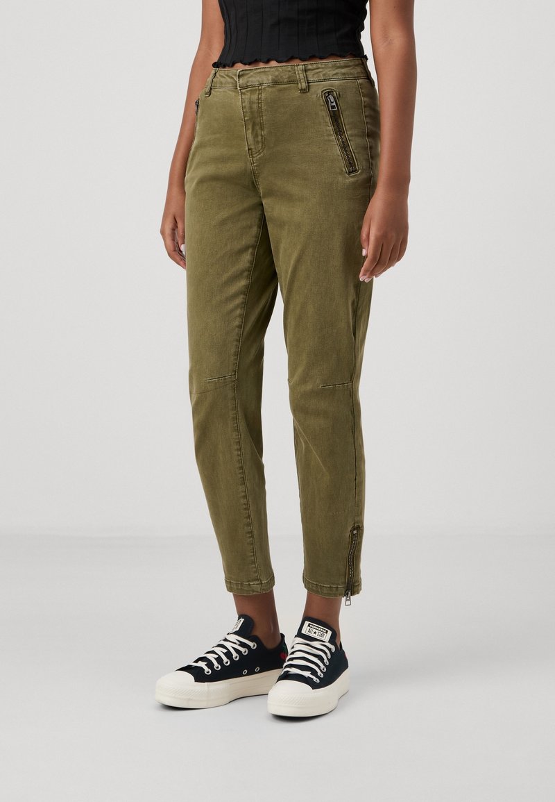 Olive green cargo pants with zippered front pockets, tapered leg, and ankle zippers. Worn with black sneakers featuring white laces.