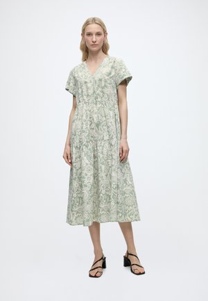 Femeie îmbrăcată într-o rochie verde de lungime midi, cu mâneci scurte și modele florale albe, purtând sandale negre cu toc, stând pe un fundal simplu.