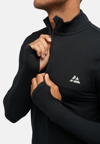 Schwarzes langärmliges Sportoberteil mit halbem Reißverschluss, Daumenlöchern und reflektierendem Logo auf der Brust. Hergestellt aus dehnbarem, glattem Stoff.