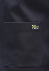 Bolsillo de tela azul marino con superficie texturizada, que presenta un logo de cocodrilo bordado en verde en la esquina superior derecha.