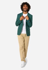 Homme souriant aux cheveux bouclés portant un blazer vert, une chemise blanche, un pantalon beige et des baskets blanches, ajustant le col de sa veste.