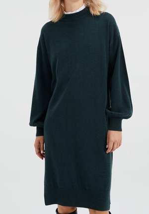 Femme portant une robe en maille manches longues vert foncé avec poignets et ourlet côtelés, et un col blanc volanté visible à l'encolure.