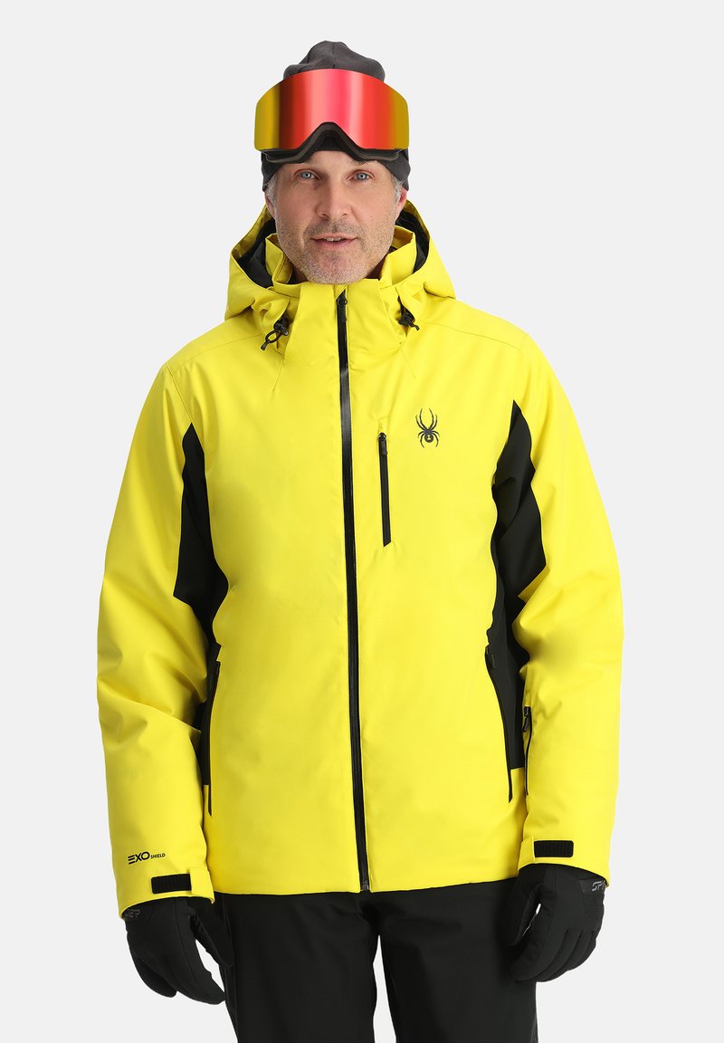 Gelbe wasserdichte Skijacke mit schwarzen Akzenten, hohem Kragen, verstellbarer Kapuze, Reißverschlusstaschen und Logo-Detail auf der Brust. Schwarze Handschuhe.
