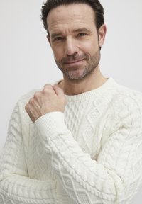 Hvit kabelstrikket genser med ribbestrikkede mansjetter og rund hals; har teksturerte mønstre og en avslappet passform. Modellen poserer med hånden på haken.