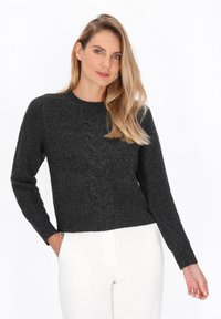Femeie cu păr lung blond, îmbrăcată cu un pulover din lână cu model în carouri de culoare gri închis și pantaloni albi, stând cu o mână în buzunar pe un fundal alb.