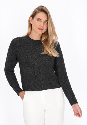 Vrouw met lang blond haar, gekleed in een donkergrijze kabeltrui en witte broek, staand met één hand in haar zak tegen een witte achtergrond.