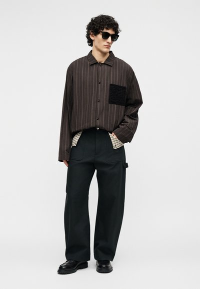 Άνδρας με σγουρά μαλλιά που φοράει μαύρα γυαλιά ηλίου, μαύρο πουκάμισο με ρίγες και τσέπη, μαύρο παντελόνι με φαρδιά μπατζάκια και μαύρα παπούτσια, μπροστά σε απλή φόντο.