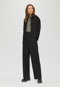 Zwarte cropped jas met frontzakken, gecombineerd met hoge zwarte broeken en een aansluitend olijfgroen topje. Gladde stof, minimalistisch ontwerp.