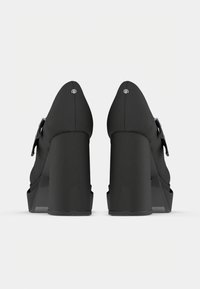 Pár černých platformových lodiček na vysokém podpatku s pásky kolem kotníku a robustními podpatky, zobrazený zezadu na jednoduchém bílém pozadí.