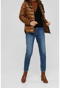 Giacca imbottita marrone con cappuccio, chiusura con zip e cordini neri, abbinata a un maglione scuro, jeans blu e stivaletti marroni con tacco.