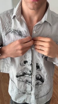 Striped button-up shirt featuring black embroidered facial design. Short sleeves, soft fabric, and white buttons. Close-fitting design.