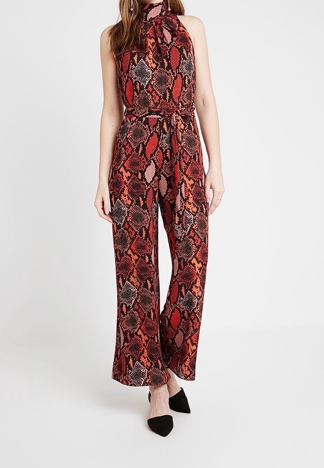 Femme portant une combinaison sans manches à col haut, motif peau de serpent rouge et noir, jambes larges et ceinture assortie, associée à des chaussures noires pointues.