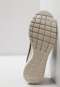 Semelle de chaussure en caoutchouc beige avec des motifs rectangulaires et carrés, reposant verticalement contre un mur blanc.
