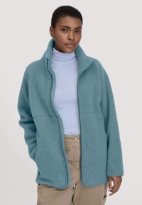 Giacca in pile azzurro chiaro con colletto alto, zip completa e due tasche frontali. Presenta una texture morbida e una vestibilità rilassata e oversize.