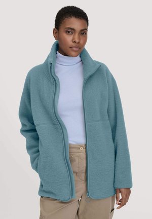 Veste en polaire bleu clair avec un col haut, fermeture éclair intégrale et deux poches avant. Elle présente une texture douce et une coupe décontractée, surdimensionnée.