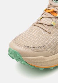 Chaussure de trail beige et vert menthe avec une semelle texturée, des lacets complexes et l'inscription "Trabuco MAX 3" près de la zone des orteils.