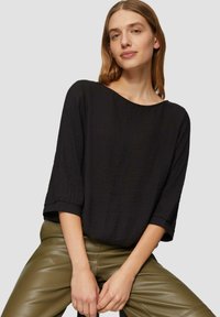 Haut noir ample à manches trois-quarts, en tissu texturé. Porté avec un pantalon en cuir vert olive, au design minimaliste.