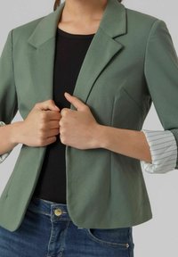 Blazer vert ajusté avec des revers pointus, présentant des manches rayées retroussées. Associé à un haut noir, les mains sont jointes au centre.