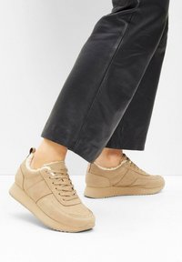 Baskets en suède beige avec une doublure en peluche, présentant un bout rond, un design à lacets plats et une semelle rembourrée. Pantalon en cuir noir visible.