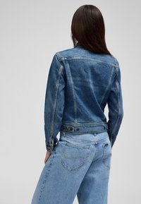 Vrouw met lang donker haar, gekleed in een getailleerd blauwe spijkerjasje en lichtblauwe spijkerbroek, staand met haar rug naar de camera.