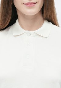 Bela polo majica iz teksturirane tkanine z značilnim klasičnim ovratnikom, trojnim zapenjanjem na gumbe in diskretno logotipom na prsih.