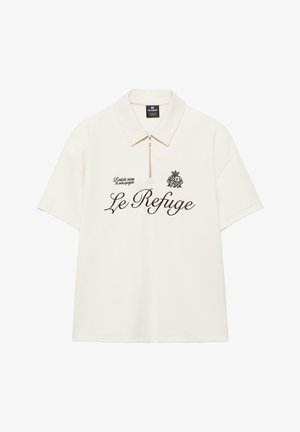 Hvid kortærmet poloshirt med lynlås, sort tekst "Le Refuge", lille våbenskjold og sætningen "L'art de vivre à son apogée" på forsiden.