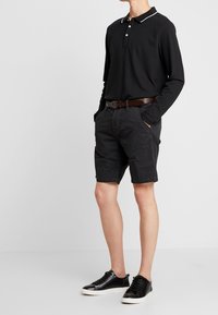 Polo negro de manga larga con collar blanco, combinado con pantalones cortos negros con estampado y zapatillas negras. Un cinturón marrón acentúa la cintura.