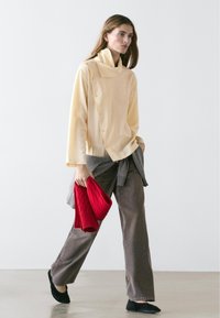 Vrouw die binnen loopt, gekleed in een beige top met lange mouwen, grijze broek met wijd uitlopende pijpen, zwarte schoenen, en een opgevouwen rode trui in haar linkerhand houdend.