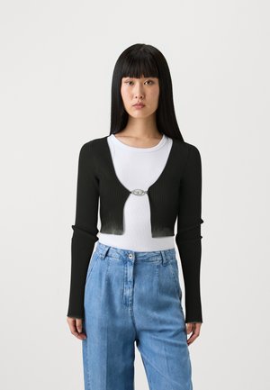 Cardigan corto a coste nero con maniche lunghe, dotato di una fibbia ovale argentata sul davanti. Indossato sopra una t-shirt bianca e pantaloni in denim blu.