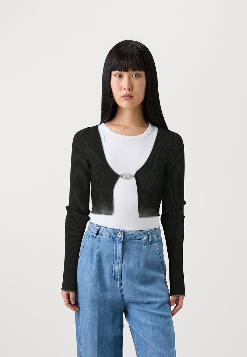 Cardigan corto a coste nero con maniche lunghe, dotato di una fibbia ovale argentata sul davanti. Indossato sopra una t-shirt bianca e pantaloni in denim blu.