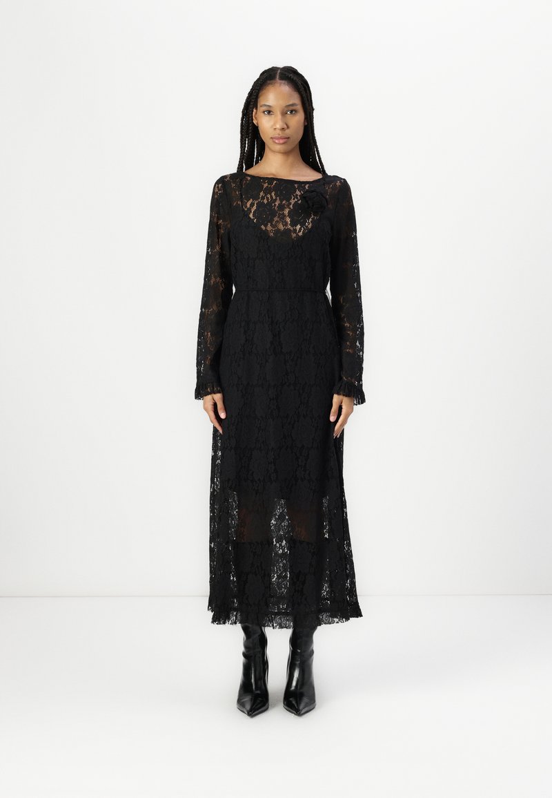 Μαύρο φόρεμα δαντέλας με μακριά μανίκια, στρογγυλή λαιμόκοψη και λουλουδάτη διάκοσμο; μέχρι τον αστράγαλο με διάφανο κατώτερο τμήμα και υφή επικάλυψη.