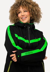 Schwarze Jacke mit leuchtend grünen horizontalen Streifen, verstellbarer Kapuze und Bündchen. Verfügt über Reißverschlüsse und Branding entlang des Ärmels.