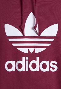 Bordeaux hoodie med en vit Adidas-logotyp med tre ränder och en ikonisk bladdesign. Dragsko vid halsen med metalldelar.