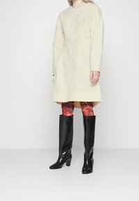 Vestido beige de gran tamaño con mangas largas, escote redondo y textura sutil, combinado con botas altas de cuero negro y leggins florales.