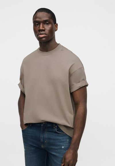 Χακί κοντομάνικη μπλούζα με γυρισμένα μανίκια, στρογγυλή λαιμόκοψη, χαλαρή εφαρμογή και διακριτική λεπτομέρεια λογότυπου κοντά στον ώμο, συνδυασμένη με σκούρα μπλε τζιν.