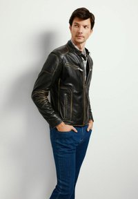 Czarna skórzana kurtka z subtelnymi brązowymi przeszyciami, z dwiema zapinanymi na zamek kieszeniami i wysokim kołnierzem, noszona na niebieskiej koszuli i dżinsach.