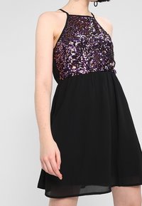 Kobieta w bezrękawnej czarnej sukience z fioletowym i czarnym cekinowym gorsetem oraz zwiewnym dołem, stojąca na tle jasnoszarym.