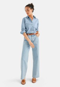 Camicia azzurra chiaro a maniche arrotolate, abbinata a jeans azzurri chiari a vita alta e una cintura marrone. Sandali a scivolo marroni completano l'outfit.