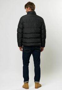 Svart pufferjakke med høy krage, horisontale sømmer og elastiske mansjetter, kombinert med mørke jeans og lysebrune boots.